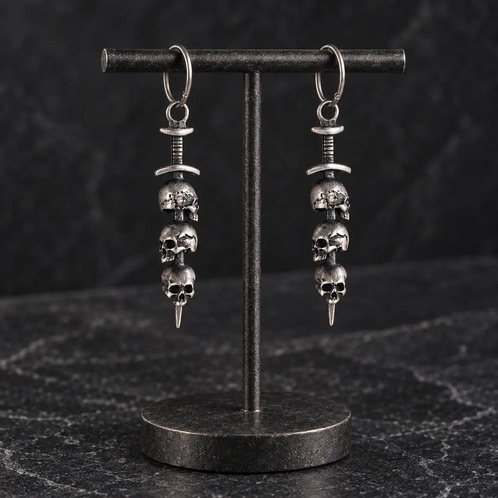 Pair of oxidized silver triple skull sword earrings hanging from a black metal jewelry display stand