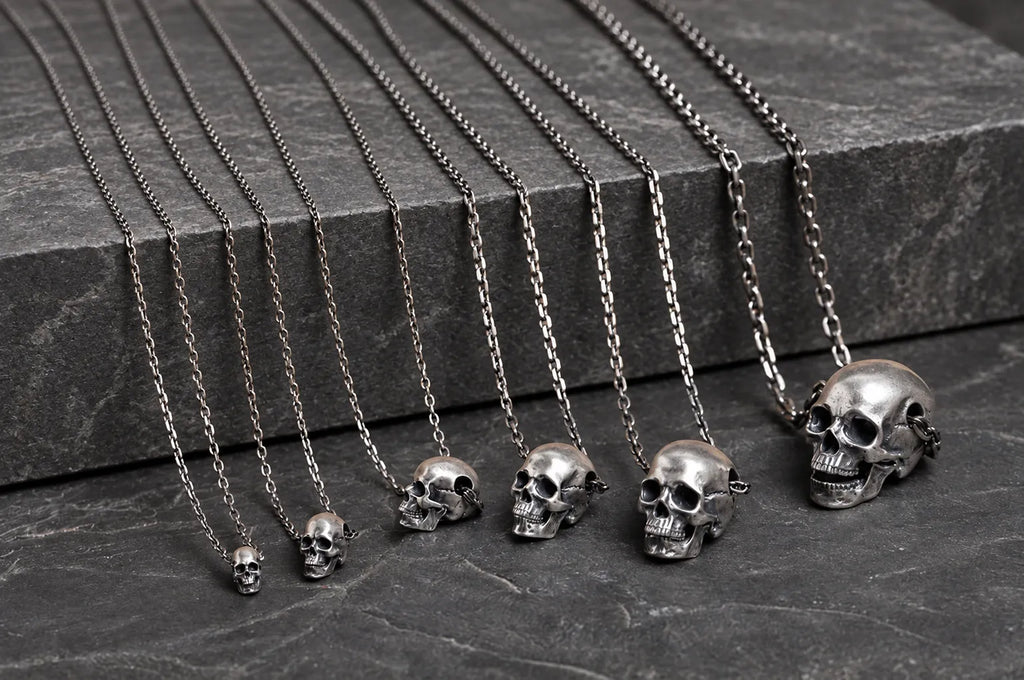 Six sizes of articulated sterling silver skull pendants displayed with naturally draped chains on dark stone