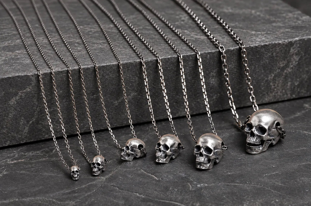 Six sizes of articulated sterling silver skull pendants displayed with naturally draped chains on dark stone