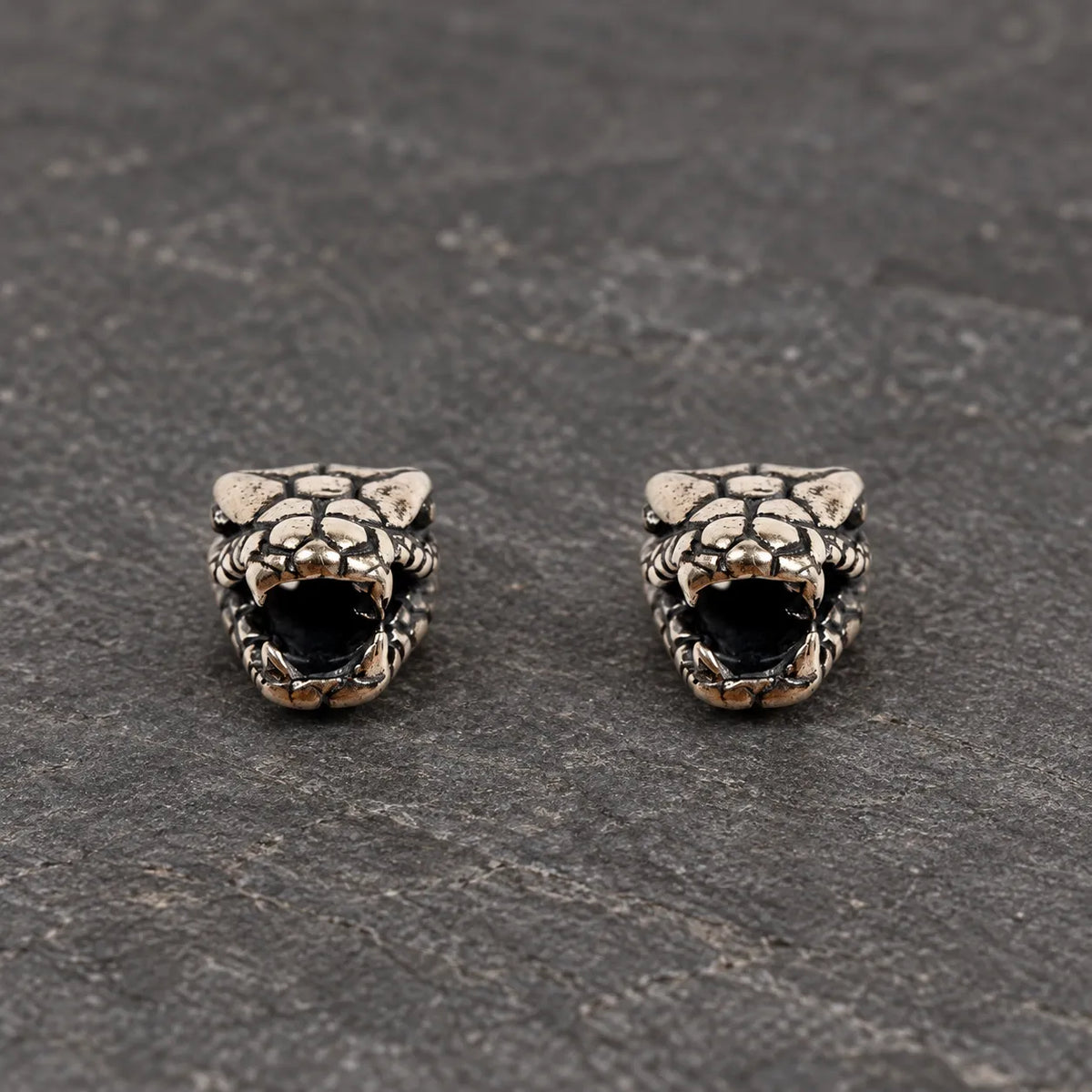 Pair of oxidized 925 sterling silver fanged serpent head stud earrings on dark stone