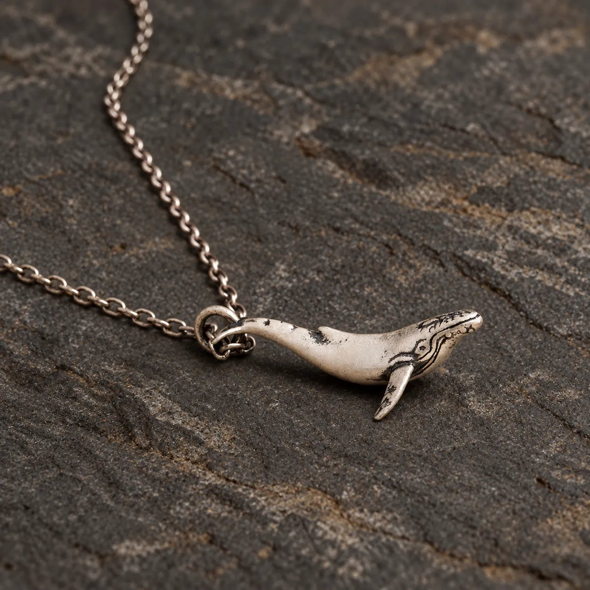 Side view of the sculptural whale pendant resting on dark stone with an optional chain