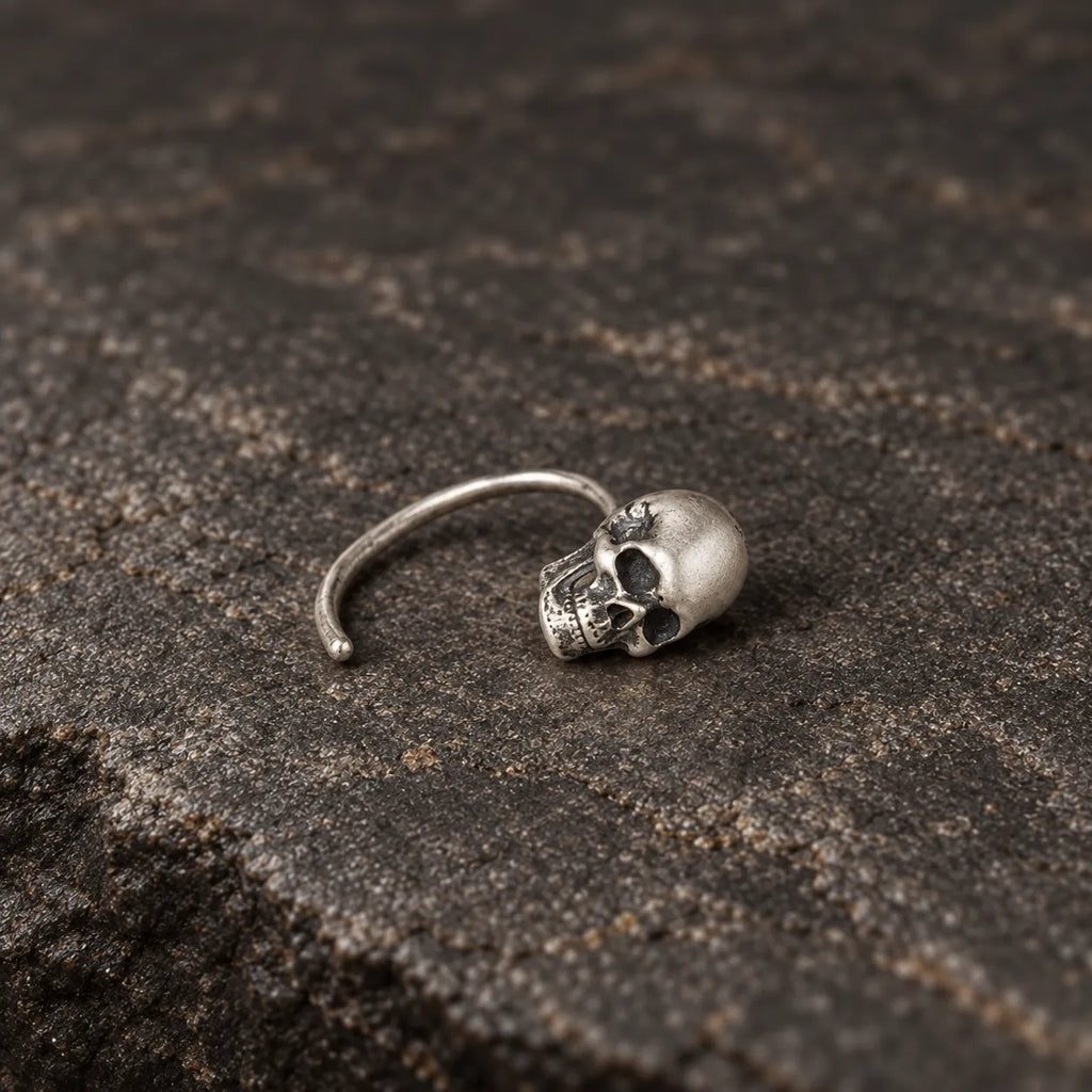 Single oxidized silver mini skull hook earring lying naturally on dark stone
