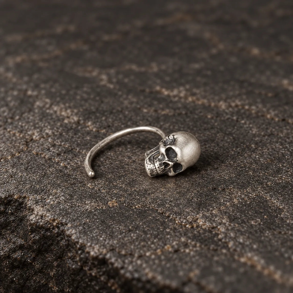 Single oxidized silver mini skull hook earring lying naturally on dark stone