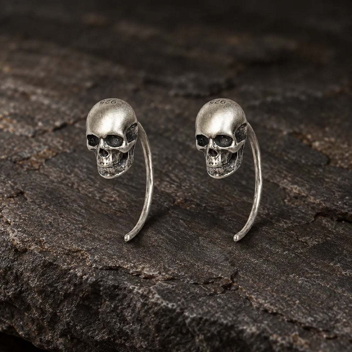 Pair of oxidized 925 sterling silver mini skull hook earrings on dark stone