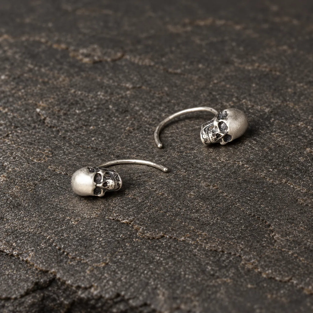Overhead view of two oxidized silver skull earrings lying naturally on dark stone