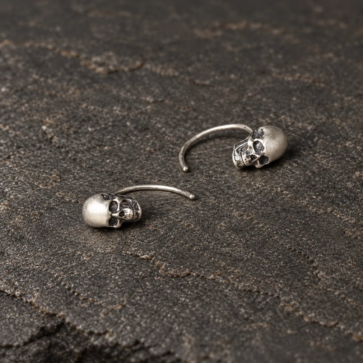 Overhead view of two oxidized silver skull earrings lying naturally on dark stone