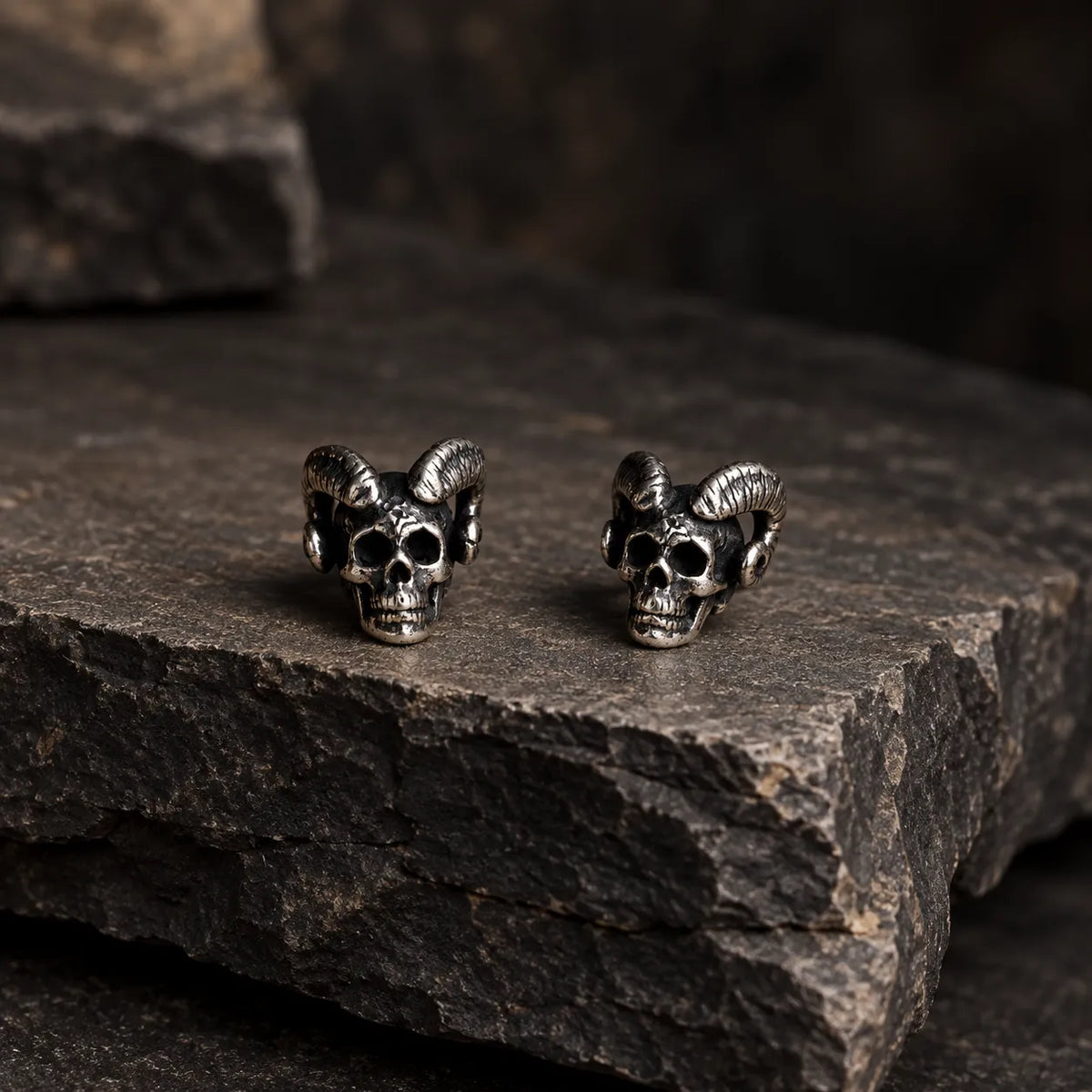 Pair of ram-horn skull stud earrings displayed on layered charcoal stone