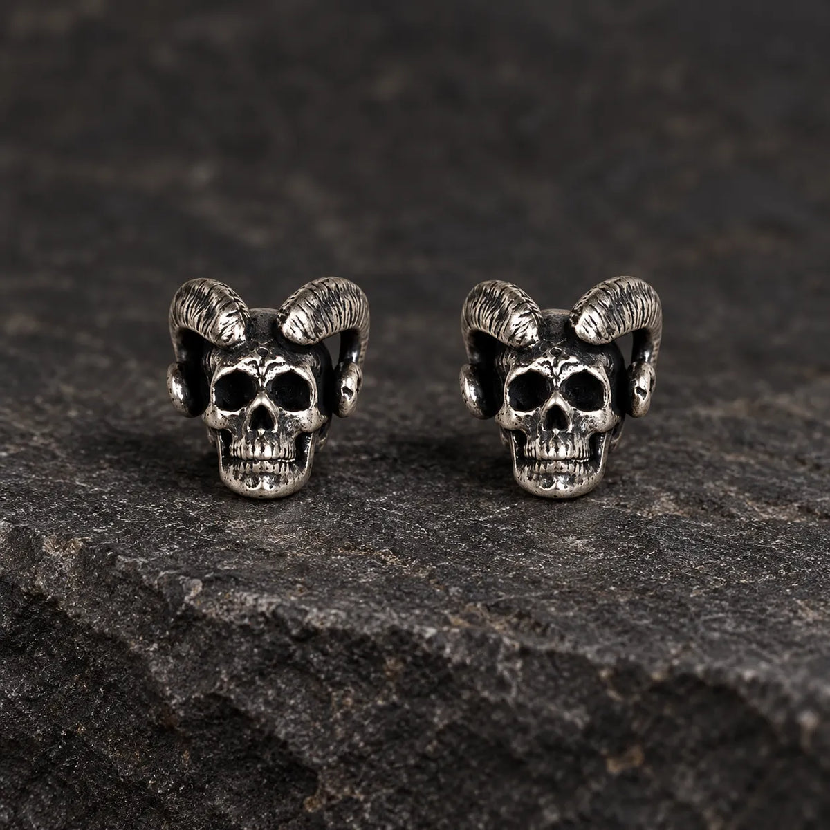 Pair of oxidized 925 sterling silver ram-horn skull stud earrings on dark stone