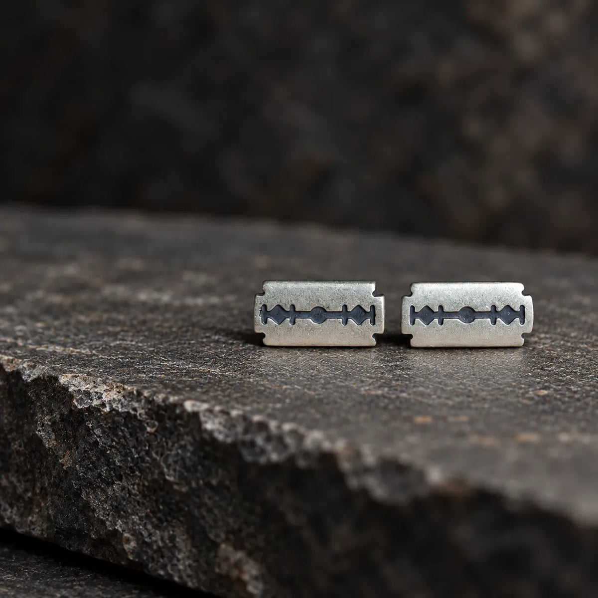 Pair of razor blade stud earrings positioned horizontally on dark stone