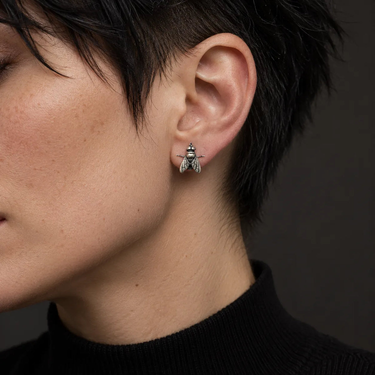 Oxidized silver fly stud earring worn at its actual 11 by 12 mm size by a short-haired model