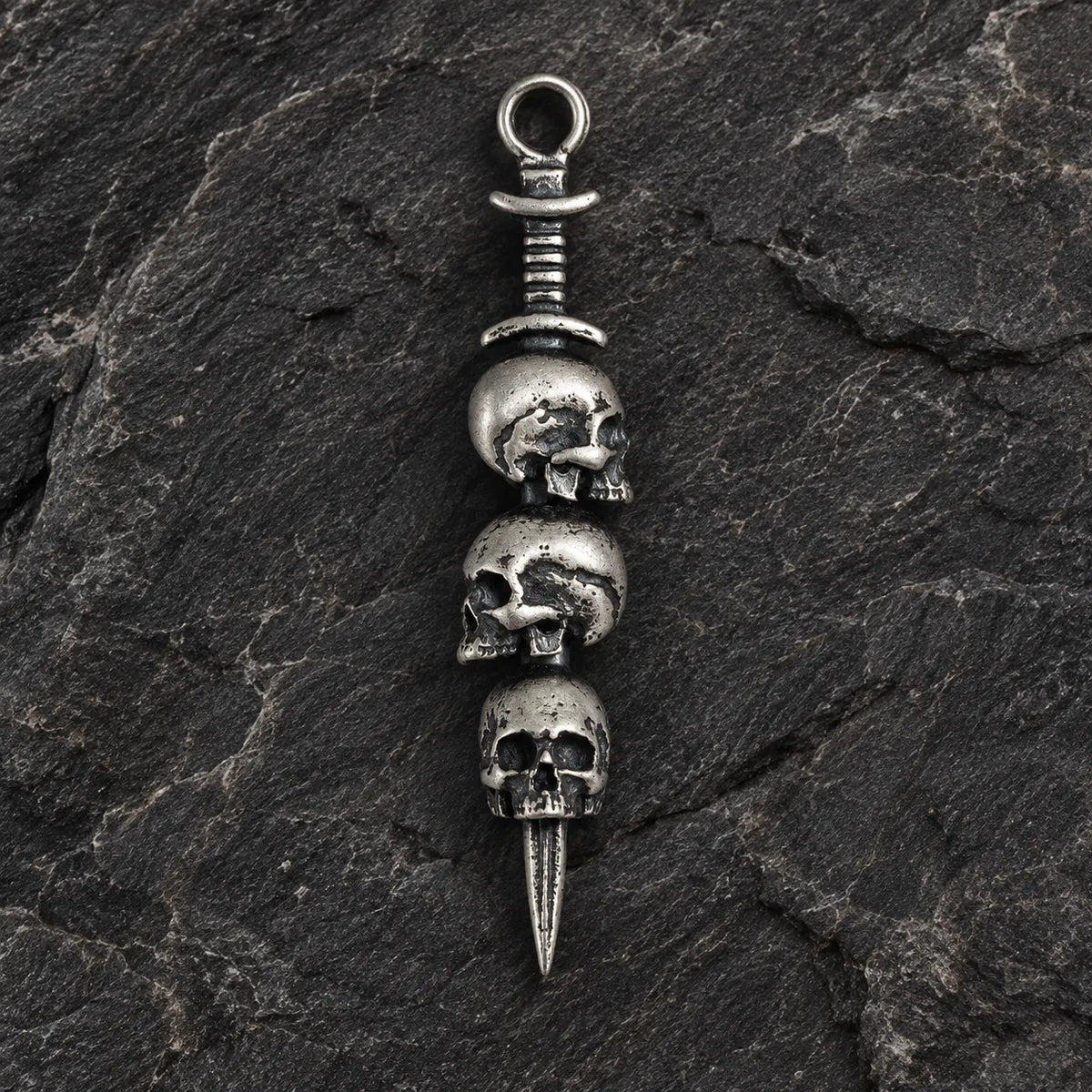 Front view of an oxidized 925 sterling silver triple skull dagger pendant on dark stone