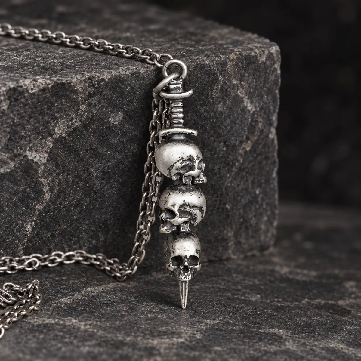 Triple skull dagger pendant hanging naturally from an optional 2.0 mm chain over dark stone