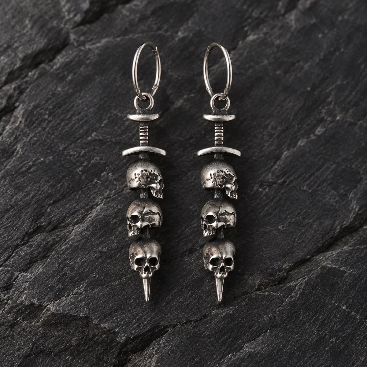 Pair of oxidized 925 sterling silver triple skull sword hoop earrings on dark stone
